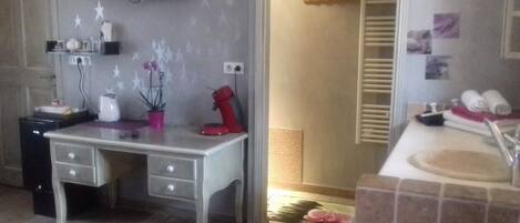 Ein Zimmer mit Schreibtisch, rotem Wasserkocher, Topfpflanze, Handtuchhalter und Duschbereich.