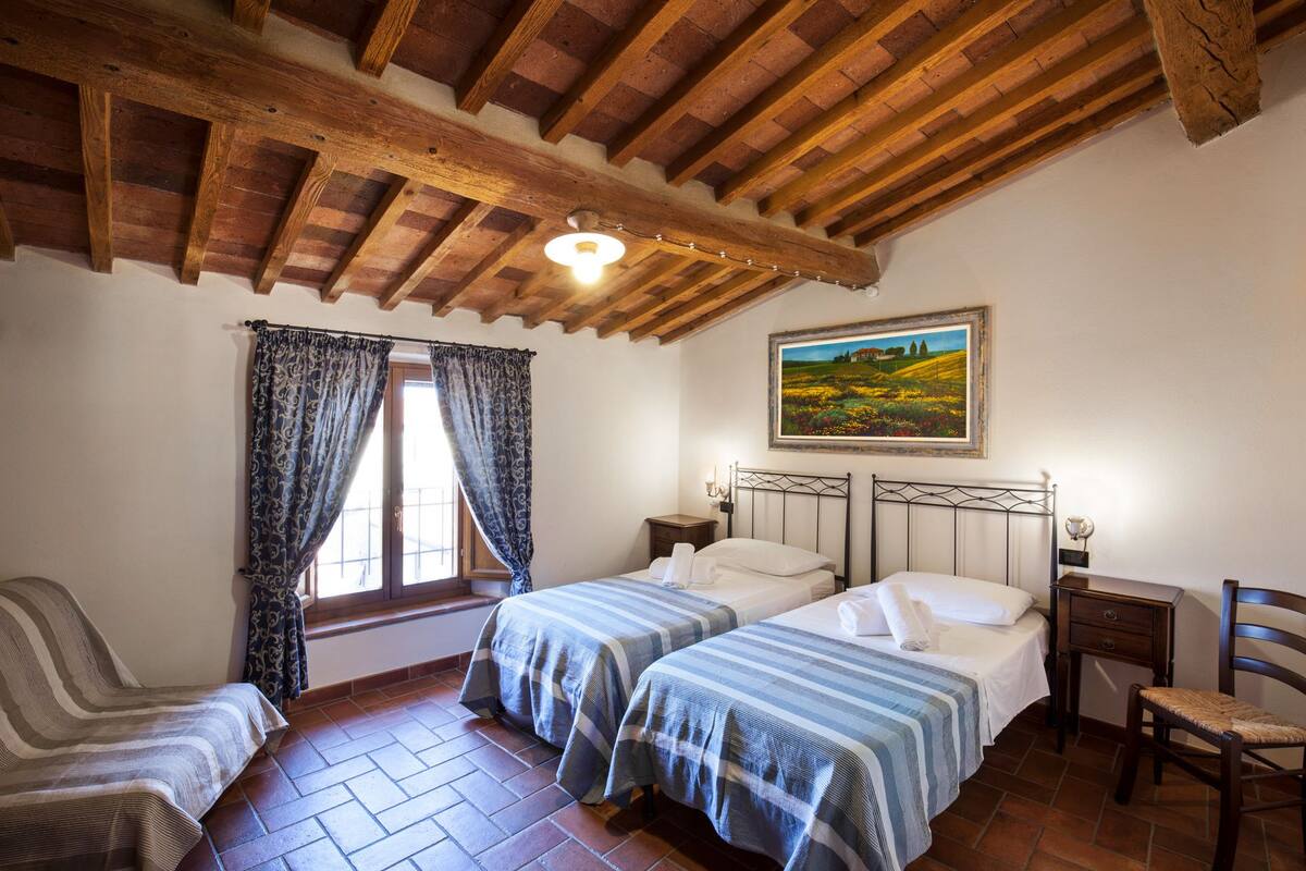 A bedroom with two beds, a wooden ceiling, a window with curtains, a painting on the wall, and a chair.