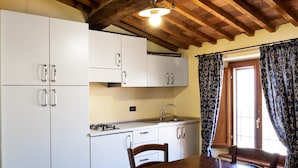 A kitchen with white cabinets, a wooden table, and a window with patterned curtains. - Residence Massa Alta (Massa Marittima)