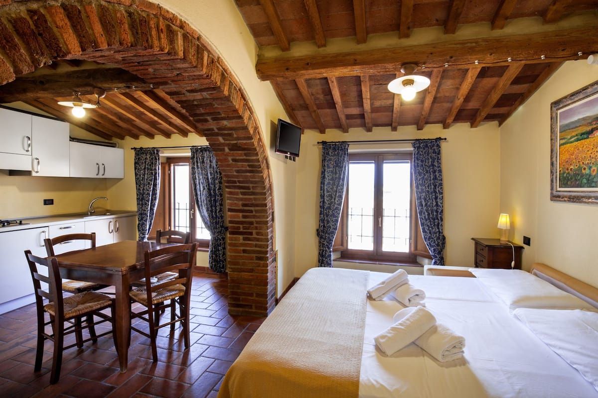 A hotel room with a wooden ceiling, a bed with white linens, a dining table with chairs, and a kitchen area with cabinets and a sink.