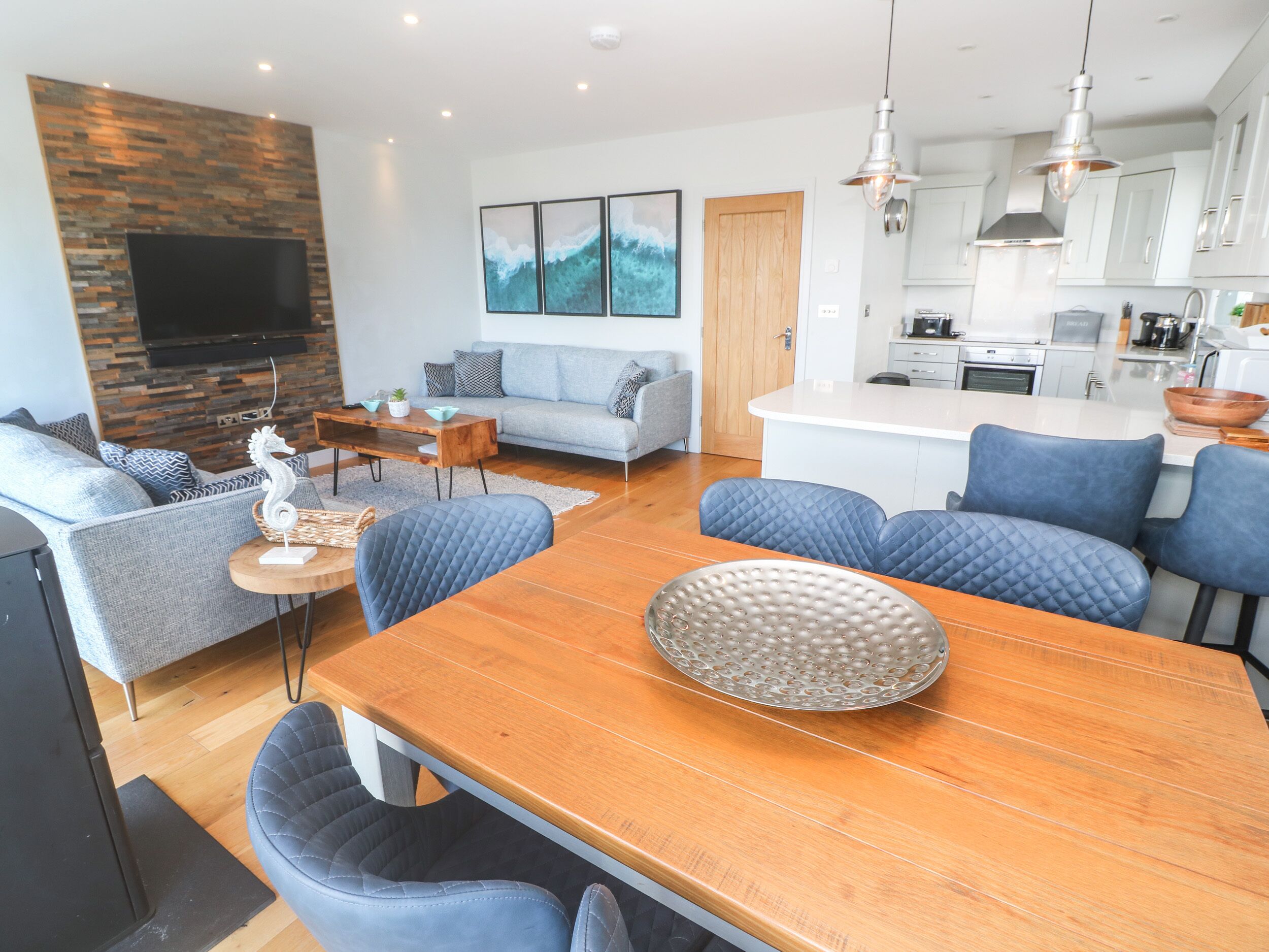 A modern living room with a wooden dining table, blue upholstered chairs, a grey sofa, and a flat-screen TV mounted on a stone wall.