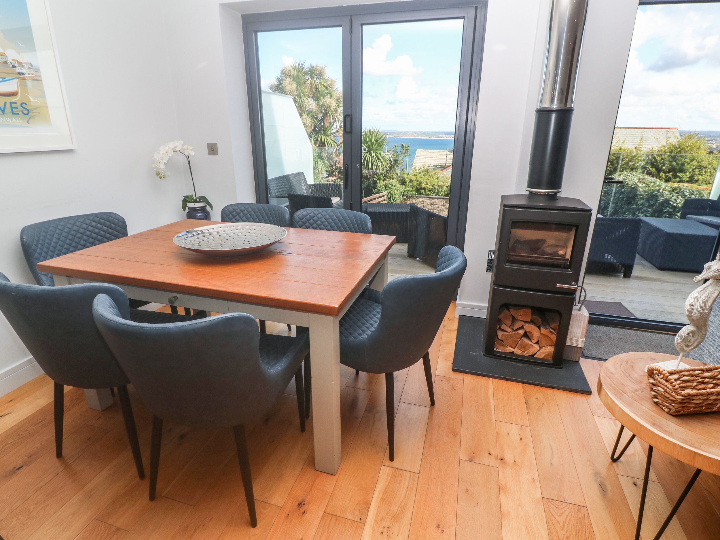 A dining area with a wooden table and chairs, a large window with a view of the sea, and a wood-burning stove.