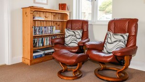 A room with two leather recliners with wooden bases, a wooden bookshelf filled with books, and a window with a view of the outside. - Wheal Hart (Newquay)