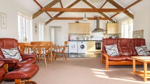 A spacious living area with a high ceiling and exposed wooden beams, a dining set, a sofa, and a kitchen in the background. - Wheal Hart (Newquay)