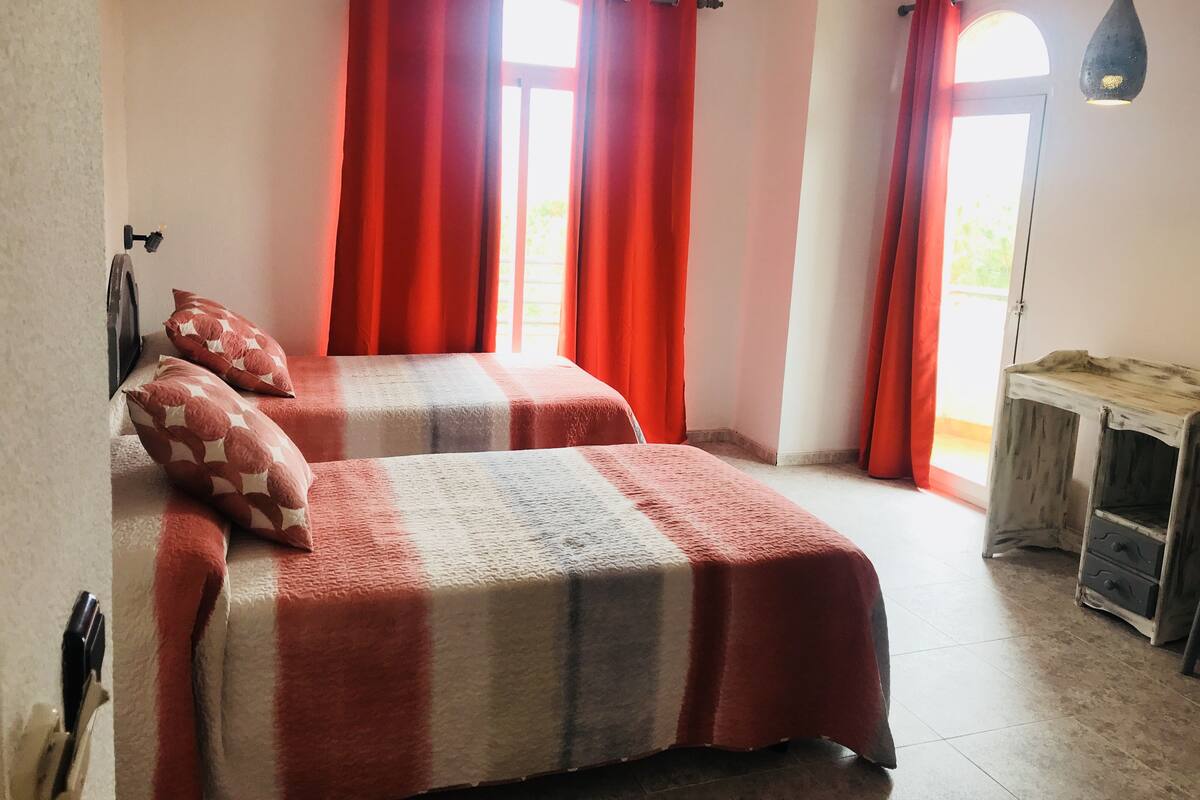 Un dormitorio con dos camas, cortinas rojas, una mesita de noche de madera y una ventana con vistas.