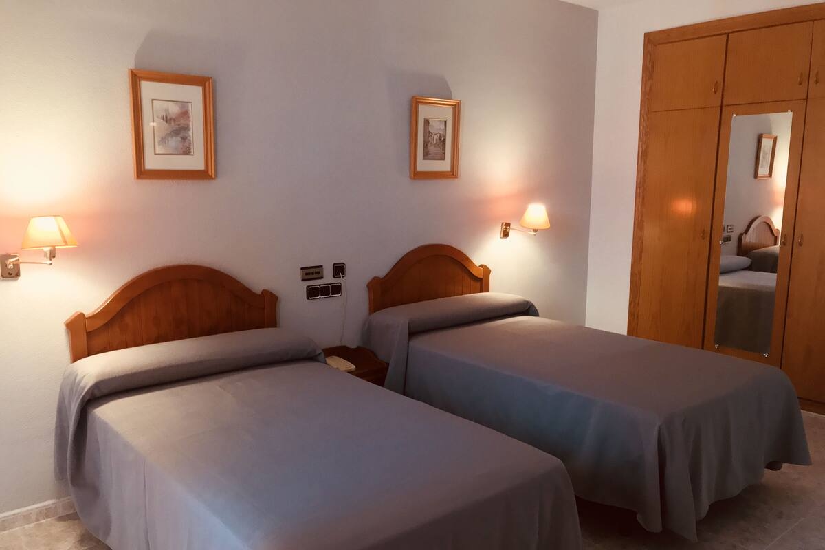 Una habitación de hotel con dos camas individuales, un armario de madera y dos lámparas fijadas a la pared.