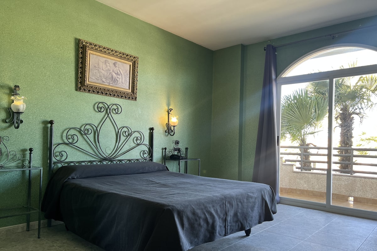 Un dormitorio con cama, paredes verdes, un cuadro enmarcado, dos apliques de pared y vistas a palmeras a través de un ventanal grande.