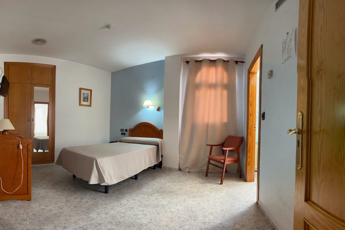 Un dormitorio con cama, una silla, un armario de madera, un escritorio con lámpara y una ventana con cortinas.