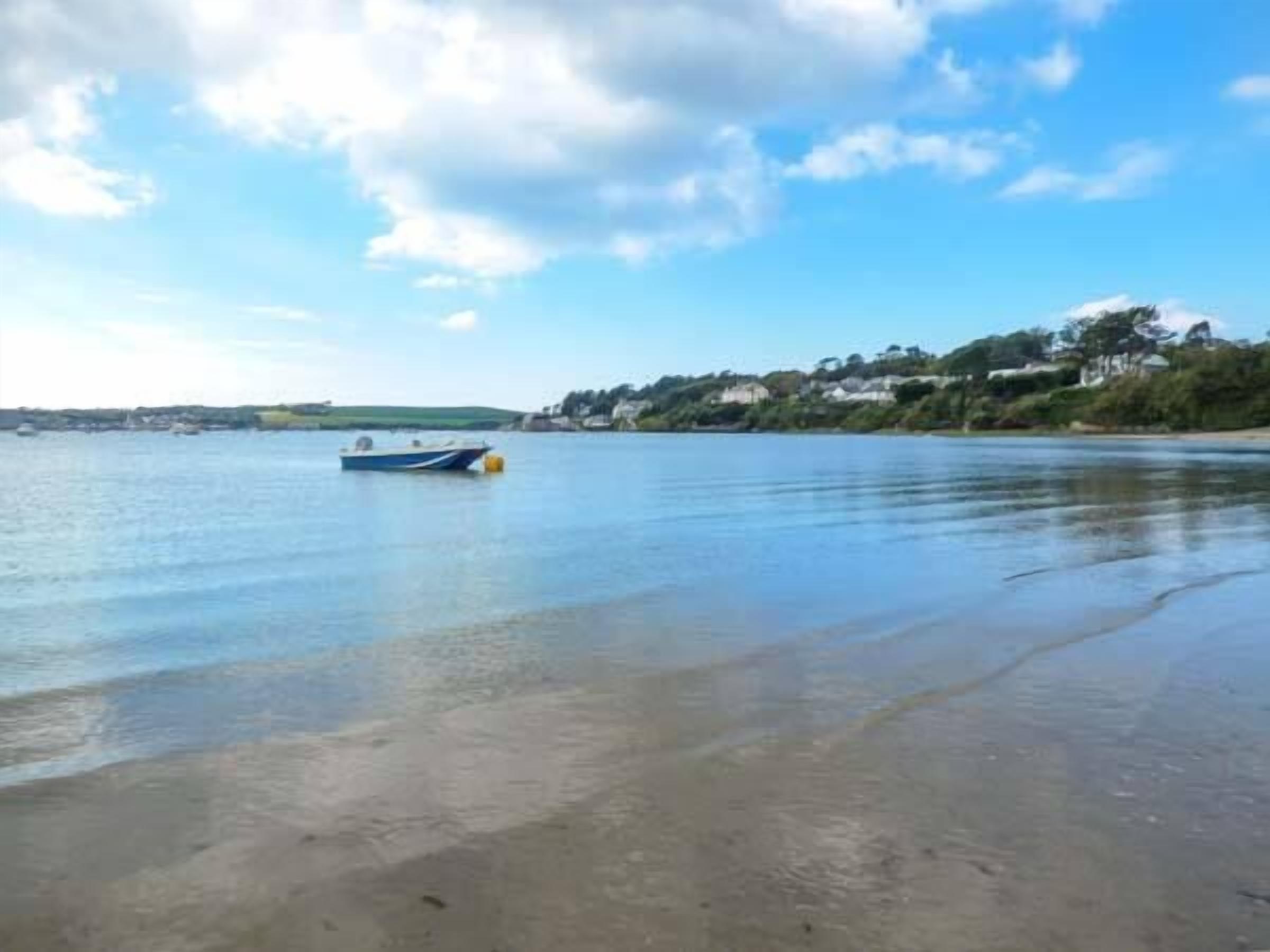 Ein Strand mit einem Boot auf ruhigem Wasser, einem sandigen Ufer und einer entfernten Küstenlinie mit Gebäuden und Grünflächen.