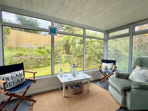 A sunroom with a view of a garden, a small table with a lantern, and two chairs. - Kilcarreg (Wadebridge)