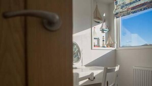 Ein Zimmer mit weißem Schreibtisch, einem Stuhl mit Sitzkissen, einem Fenster mit Blick zum Himmel und einer Heizung.