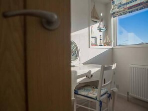 A room with a wooden door, a white desk, a chair with a cushion, a window with a view of the sky, and a radiator. - Kilcarreg (Wadebridge)