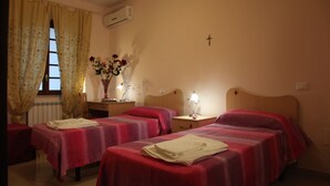 Habitación de hotel con dos camas individuales, una mesita con lámpara, un florero con flores y una cruz en la pared.