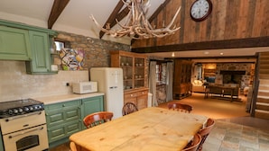 A rustic kitchen with a stone wall, wooden beams, a large dining table, and a clock on the wall. - Barn Cottage (Hayle)