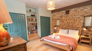 A bedroom with a stone wall, a wooden bed with a woven headboard, a bookshelf, and a lamp on a nightstand. - Barn Cottage (Hayle)