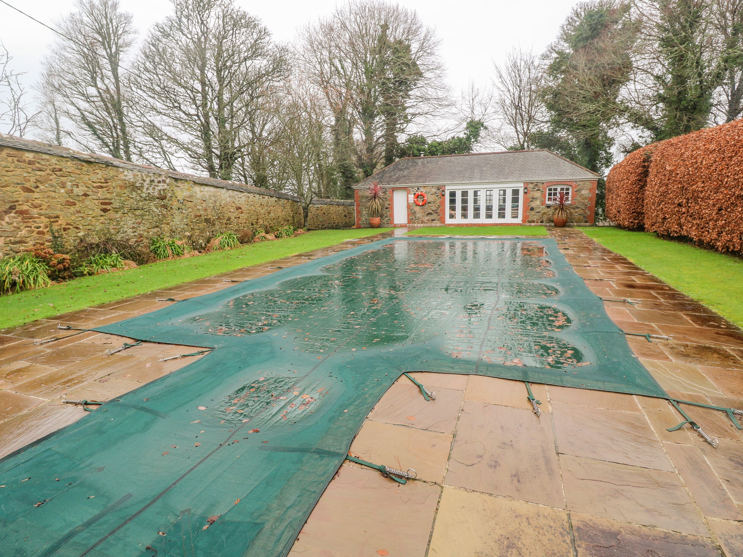 A swimming pool with a green cover, surrounded by a stone wall and a hedge.