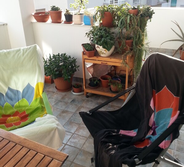 A patio with a wooden chair and a stroller, surrounded by potted plants and a small wooden shelf.