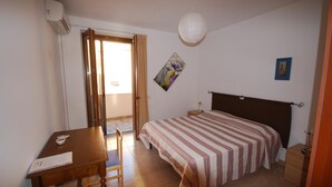 A bedroom with a bed, a desk, and a chair. There is a window with curtains, a painting on the wall, and a small bedside table.