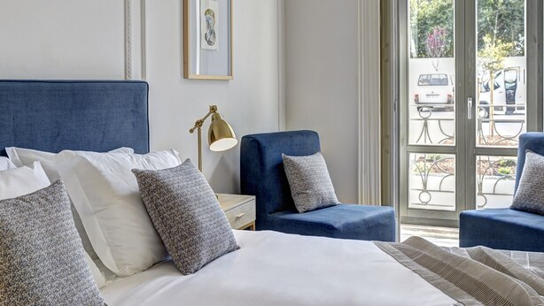 A hotel room with a large bed, two blue armchairs, a bedside table with a lamp, and a framed picture on the wall.