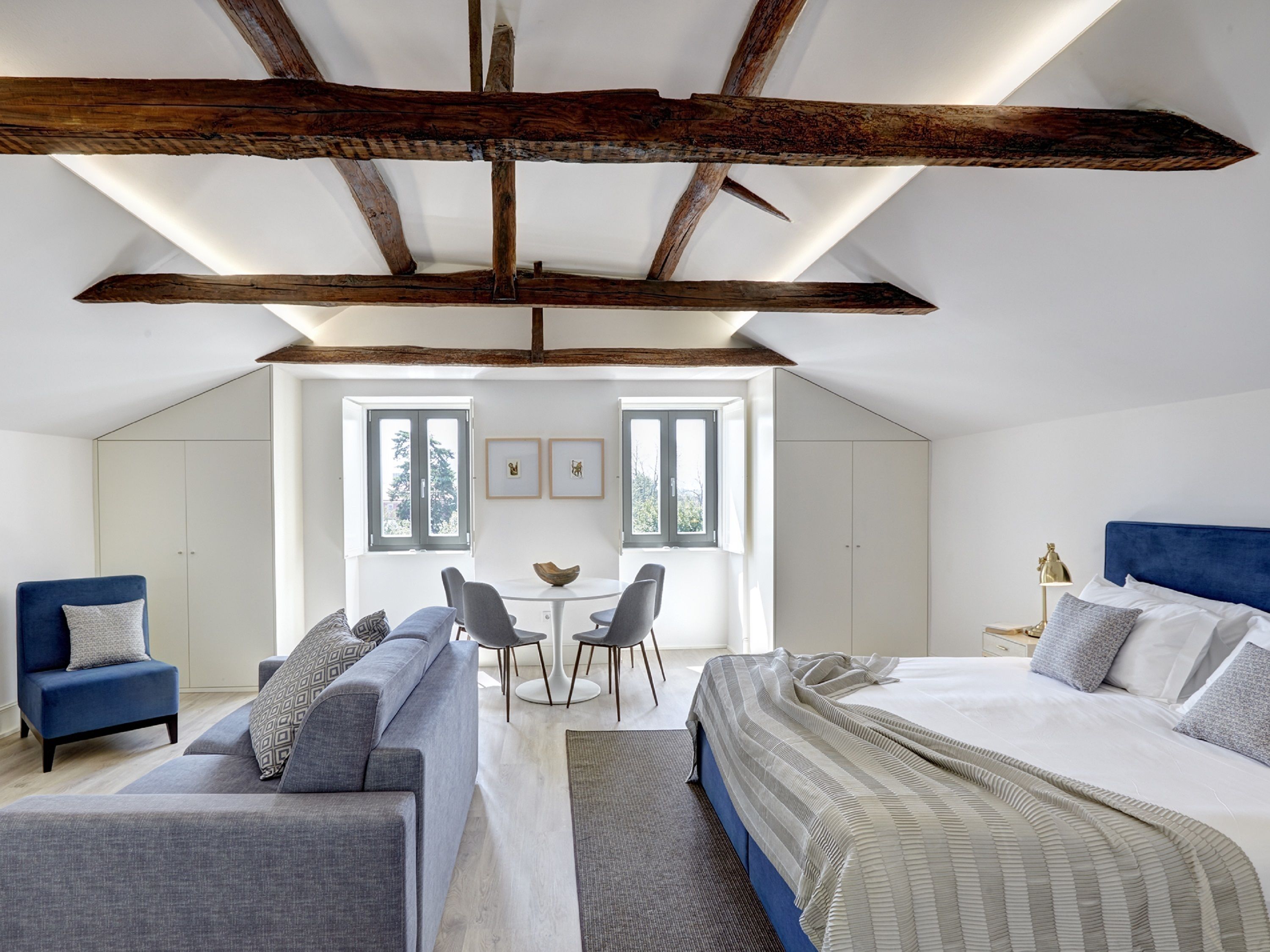 A modern bedroom with a loft-style ceiling, a bed, a sofa, a small dining table, and two chairs. There are windows providing natural light and a view of the outdoors.