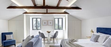 A modern bedroom with a loft-style ceiling, a bed, a sofa, a small dining table, and two chairs. There are windows providing natural light and a view of the outdoors.