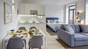 A modern hotel room with a dining area, kitchenette, and a bed with a blue headboard.