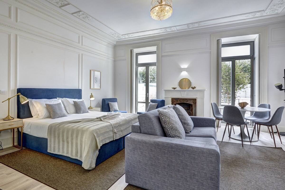 Une chambre spacieuse avec un grand lit, un coin salon avec un canapé et des fauteuils, une cheminée et de grandes fenêtres donnant sur l’extérieur.