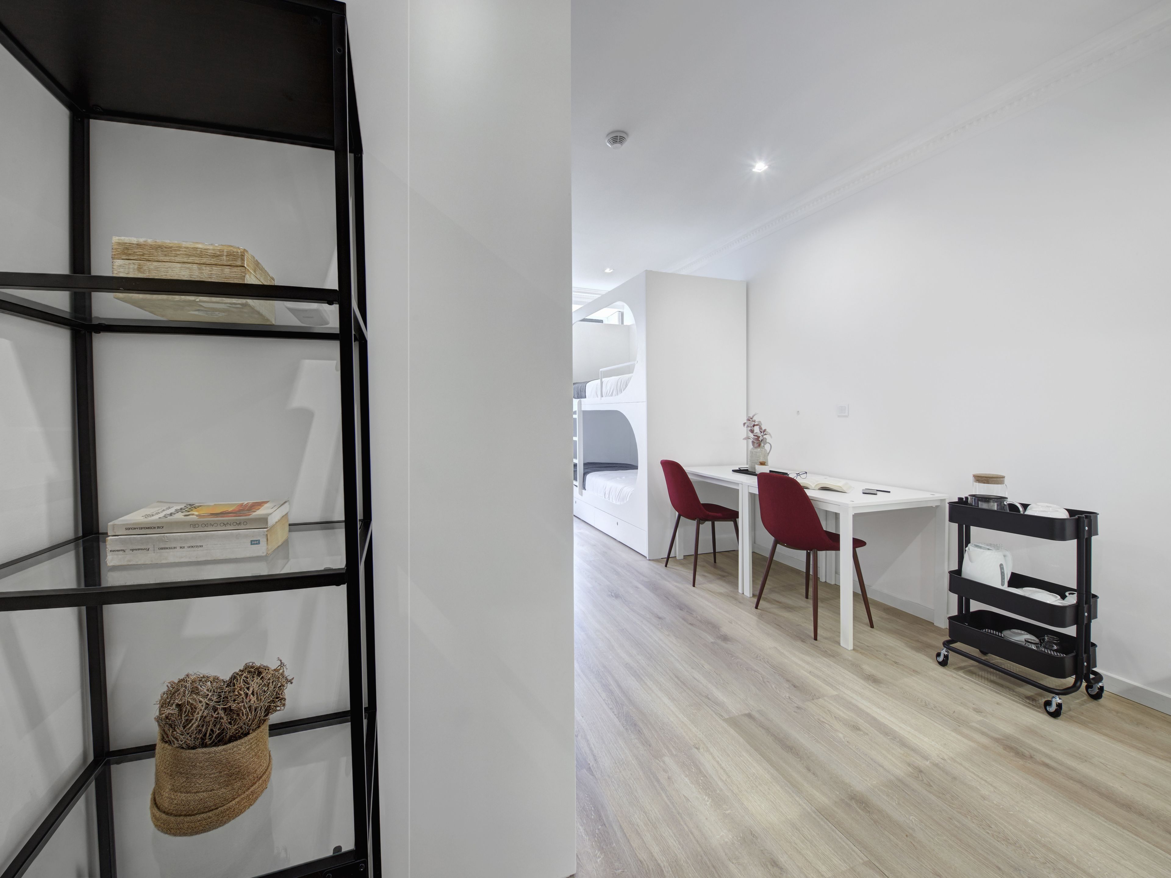 A modern room with a white desk, red chairs, a black shelving unit, and a white bunk bed in the background.