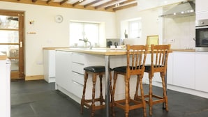 A modern kitchen with wooden beams, white cabinets, a stainless steel oven, and a bar area with stools. - Eastcott Farmhouse (Holsworthy)