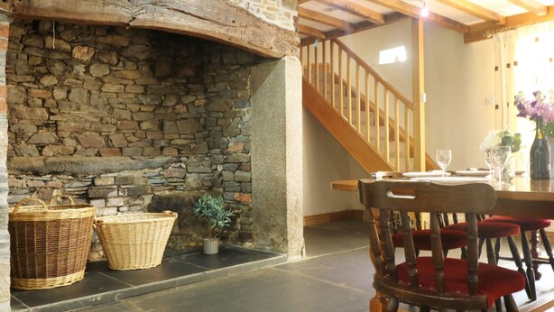 A stone fireplace with baskets, a wooden staircase, and a dining area with chairs and a table. - Eastcott Farmhouse (Holsworthy)