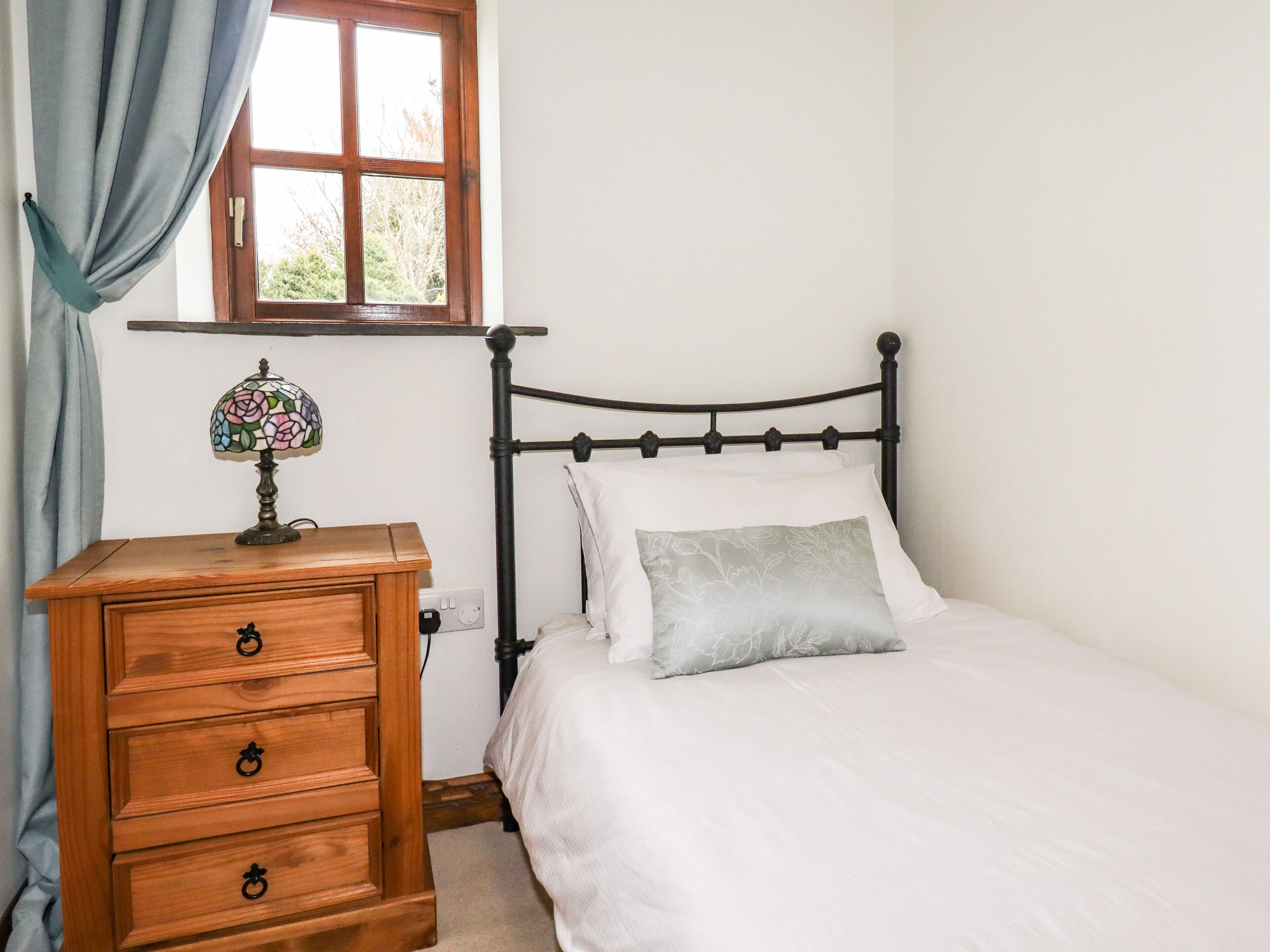 A bedroom with a wooden nightstand, a bed with white bedding, a floral lamp, and a window with curtains.