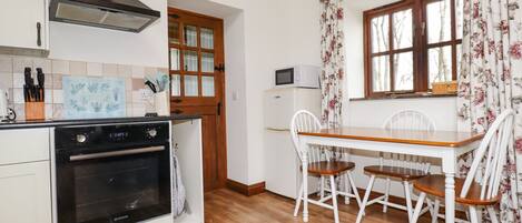 A compact kitchen with a stove, microwave, and wooden cabinets. There's a dining table with chairs and a window with floral curtains.
