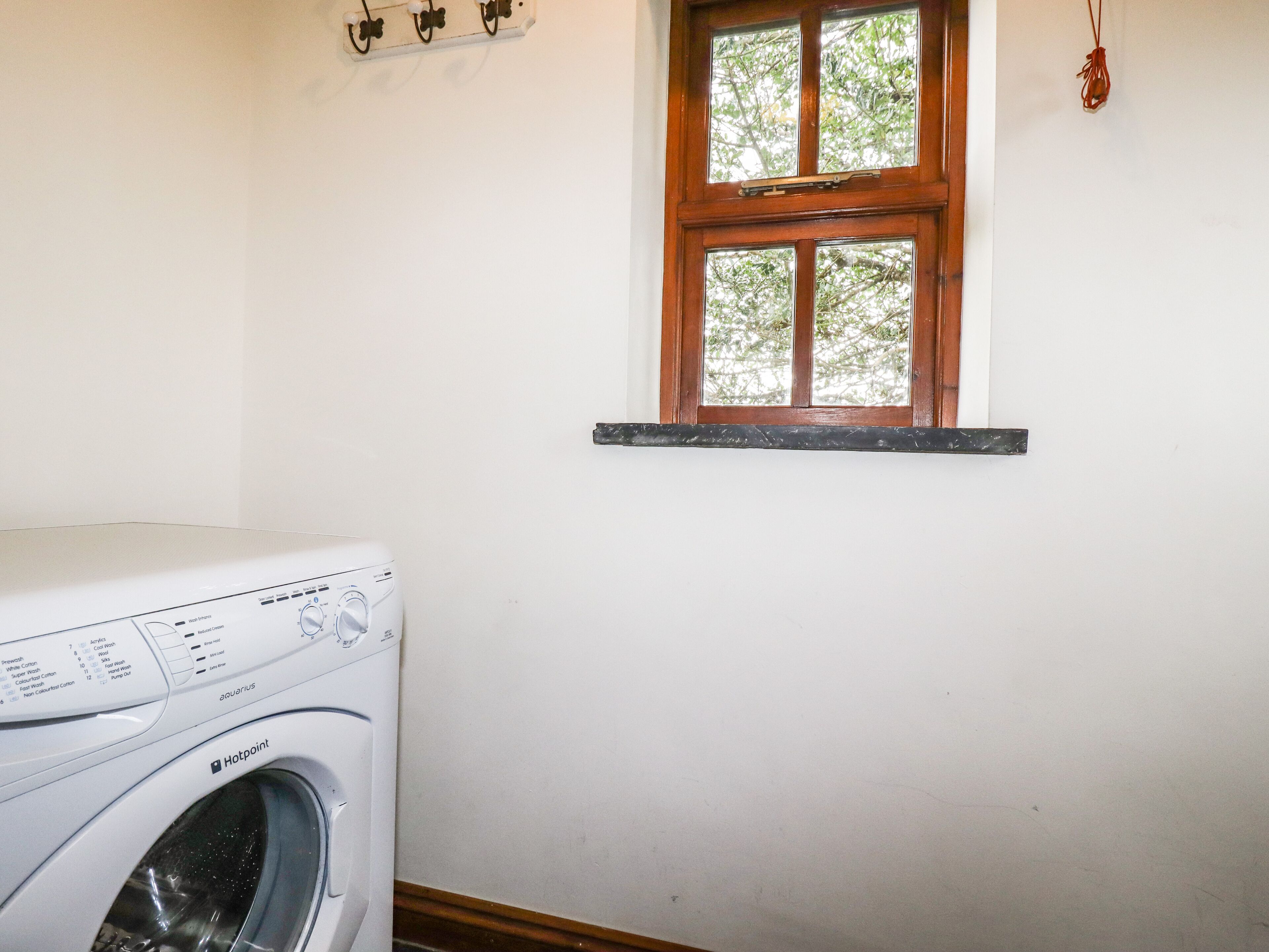 A laundry room with a white washing machine, a window with a wooden frame, and a wall-mounted shelf with hooks.