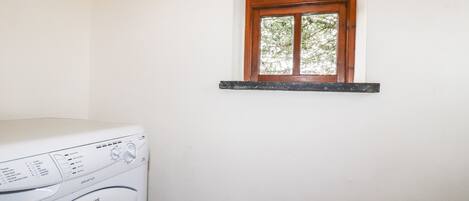 A laundry room with a white washing machine, a window with a wooden frame, and a wall-mounted shelf with hooks.