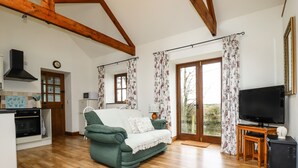 A cozy living room with a green sofa, wooden beams, a television, and a kitchen area with a microwave. - Brocks Barn (Bodmin)