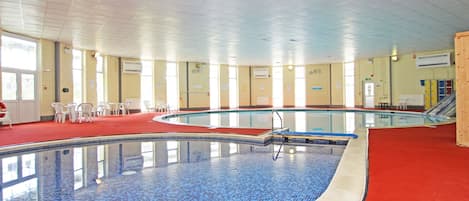 An indoor swimming pool with clear blue tiles, surrounded by white chairs and a red carpeted area.