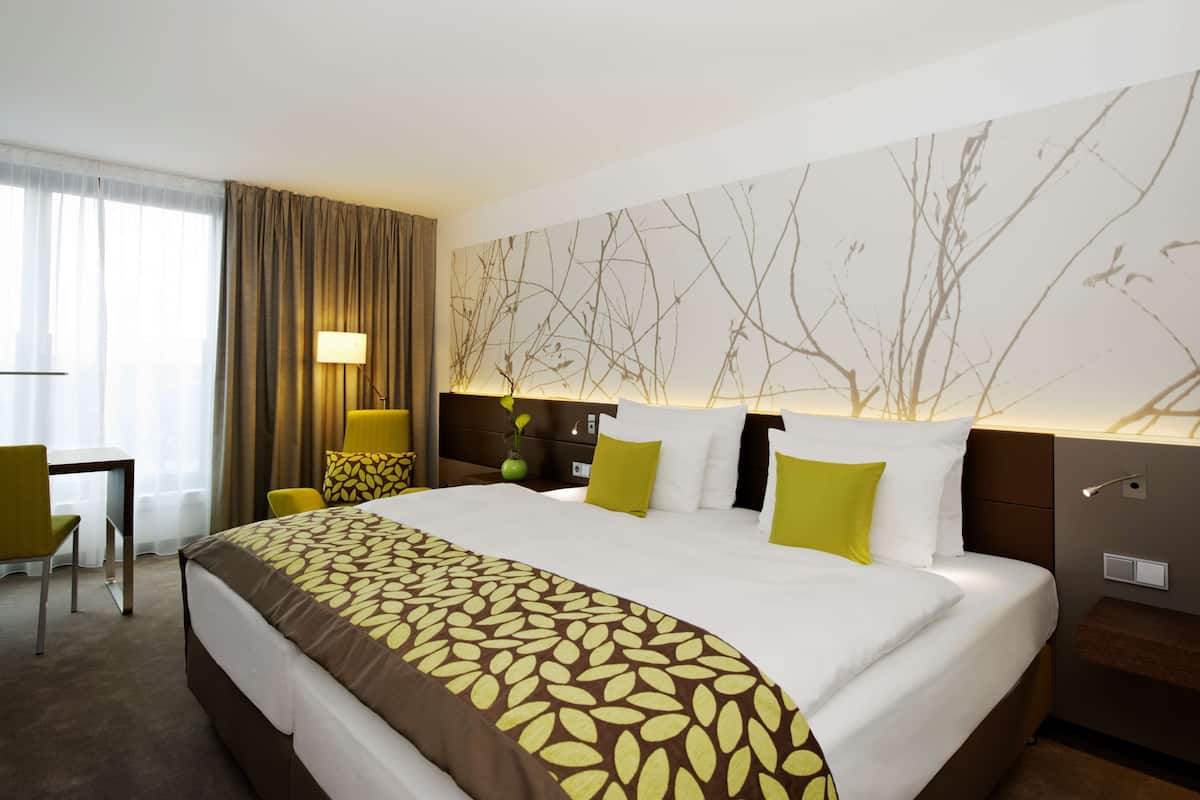 A hotel room with a large bed, a desk, a chair, a lamp, and a wall with a tree branch design.