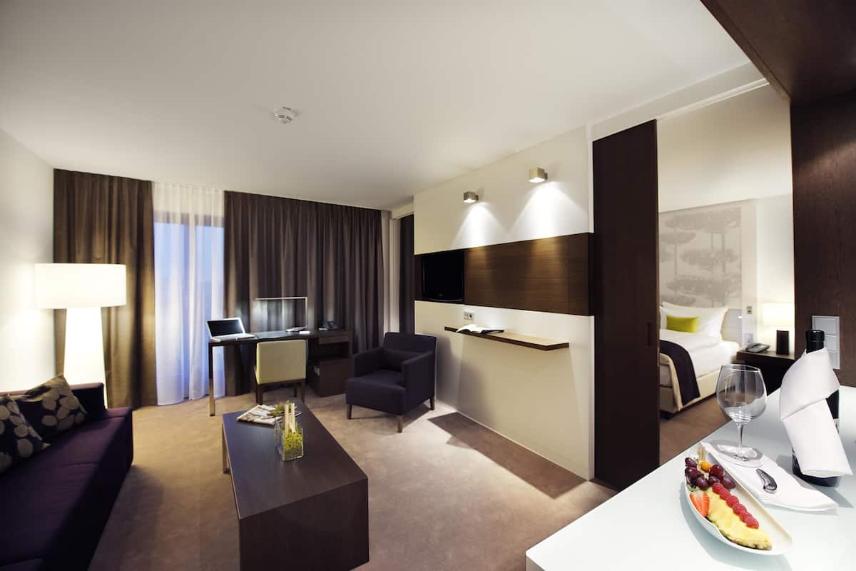 A modern hotel room with a sofa, a desk with a laptop, a dining area with a plate of fruit, and a bed with a nightstand.