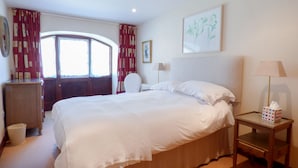 A hotel room with a large bed, a bedside table with a lamp, a chair, a window with curtains, and a painting on the wall. - The Granary (Hayle)