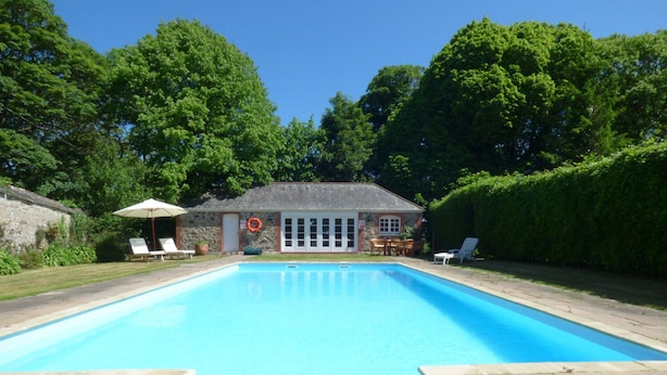 A rectangular swimming pool with a stone deck, surrounded by a stone wall and a hedge. There is a small building with a door and windows, and a lifebuoy hanging on the wall. - The Granary (Hayle)