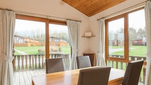 A room with a wooden table and chairs, large windows with curtains, a small shelf with a lamp, and a view of a grassy area with buildings. - High Tide (Wadebridge)