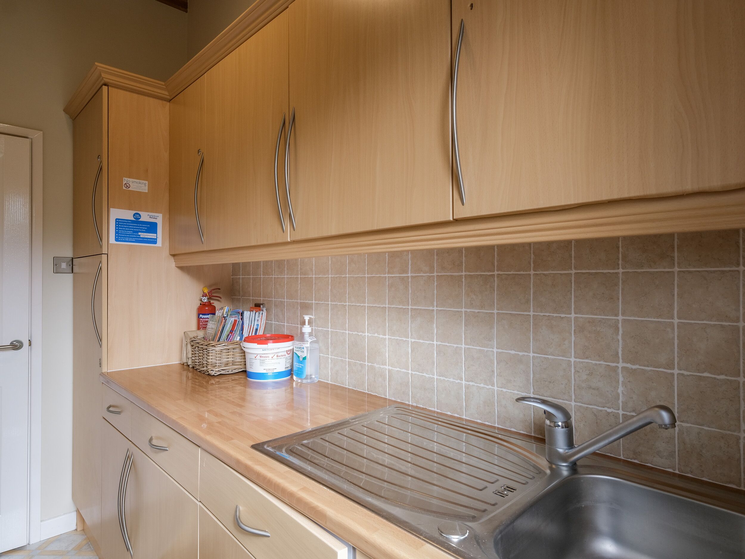 A kitchen with wooden cabinets, a stainless steel sink, a tiled backsplash, and a countertop with a kettle, a container, and a bottle of hand soap.
