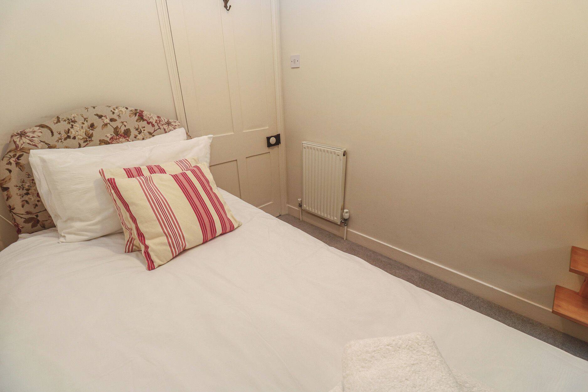 A neatly made bed with white bedding and two pillows, one striped and one plain, in a room with a radiator and a wooden shelf.