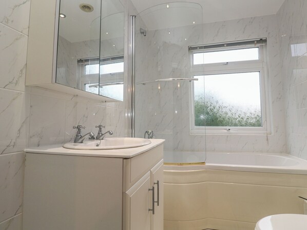 A modern bathroom with a marble-tiled wall, a large mirror, a sink with two faucets, a bathtub, and a window with a view of trees. - Banneth (Helston)