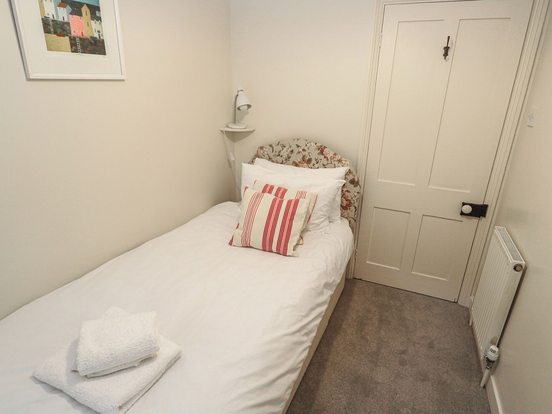 Een eenpersoonsbed met witte beddengoed en gestreepte kussens, een bedlamp, een radiator en een deur met een deurklink.