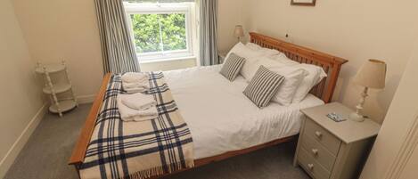 A neatly made bed with a plaid blanket and folded towels, a bedside lamp, a small bedside table, and a window with curtains.
