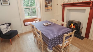Une salle à manger avec une table dressée pour quatre personnes, une cheminée, une horloge et une fenêtre donnant sur un paysage verdoyant.