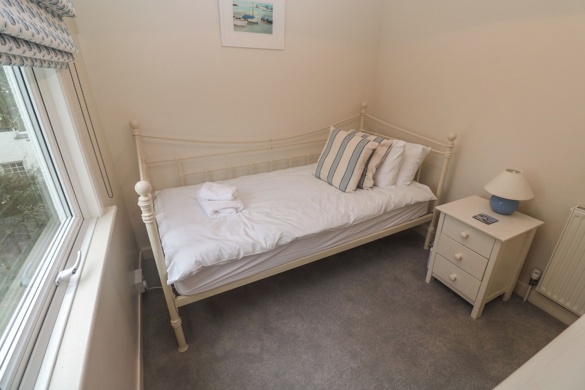 A small, single-bed room with a white bed, a bedside table with a lamp, and a window with blinds.