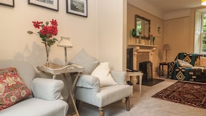 Un salon avec un canapé, un fauteuil et une petite table sur laquelle se trouve une lampe. On aperçoit un foyer au fond de la pièce et une fenêtre donnant sur un paysage verdoyant.