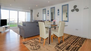 Ein modernes Wohnzimmer mit grauem Sofa, einem Glastisch mit weißen Stühlen, einem Flachbildfernseher und einem großen Fenster mit Blick nach draußen.